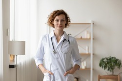 Confident young female physician standing in medical office. Proud professional woman doctor therapist looking at camera. Portrait of lady general practitioner wearing white coat and stethoscope.