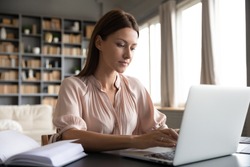Focused woman journalist freelancer working online on laptop, sitting at desk at home, looking at screen, typing, serious young female writing blog or chatting with friends in social network