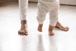 Crop close up of small biracial toddler infant make first steps on home wooden floor holding mom hands, little african American baby child learn walking with mother support care, childcare concept