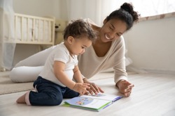 Smiling young african American mother sit on warm floor play with little infant toddler child, happy biracial mom relax have fun read book with small baby girl at home, motherhood, childcare concept