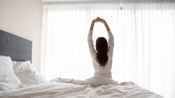 Rear view woman wake up sit in bed raised hands stretching body start day right with exercises, increases muscles tone encouraging joints flexibility, energy boost, enjoy new day, good morning concept
