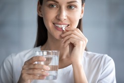 Close up image young adult beautiful 35s woman smiles takes pill holding glass of water, concept of minerals and vitamins for female, hair skin nail care dietary supplement, daily dosage of medication