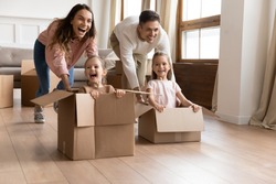 Happy parents playing with children in new apartment living room, two cute little daughters sitting in cardboard boxes, laughing mother and father pushing, family celebrating moving day