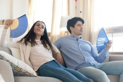 Exhausted young man and woman sit on couch at home breathe fresh air form waver, lack air conditioner, overheated couple rest on sofa suffer from heatstroke or hot weather, wave with hand fan