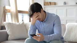 Upset young man sit on sofa at home frustrated by negative message on smartphone gadget, unhappy male distressed by eviction notice email or bad news on cellphone, get cell problems or virus