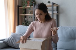 Angry confused woman unpacking parcel, wrong or broken online store order, sitting on couch at home, dissatisfied female looking in cardboard box, bad delivery service, displeased by post shipping