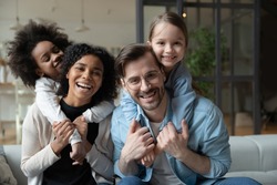 Cheery African and Caucasian girls cuddle piggy back multi-racial parents loving mom and dad seated on sofa in living room. Happy multinational family portrait, new home funny weekend together concept