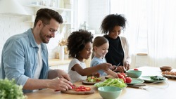 Full multinational family with cute daughters preparing dietary meal natural nutrition, cutting fresh vegetable for salad, parents caring for children health eat organic food, weekend activity at home
