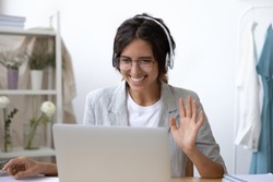 Smiling young Caucasian woman in headset wave greet talking on webcam virtual conversation on laptop, happy female in wireless headphones speak on video call on computer, consult client online