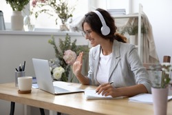Smiling young Caucasian woman in headphones take online educational course or training on laptop from home, happy female in wireless headset wave to camera, talk on webcam video call on computer