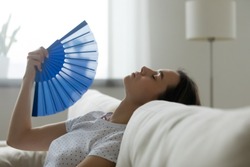 Close up image woman puts head on sofa cushions closed eyes feels sluggish due unbearable heat, waves hand blue fan cool herself, hot summer flat without air-conditioner climate control system concept