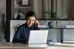 Smiling Indian businesswoman feels satisfied confident provide help consulting client distantly by mobile phone call seated at desk in modern office. Busy fruitful workday, successful employee concept