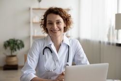 Portrait of smiling young Caucasian woman nurse sit at desk in hospital or private clinic, profile picture of happy positive female doctor GP in white medical uniform at workplace, healthcare concept