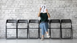 On grey brick wall background anonymous young woman sit on chair hiding her face behind paper with interrogation symbol. Doubtful female holding question mark sheet, problems and solutions HR concept
