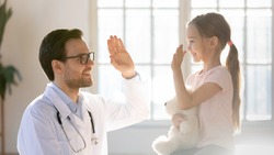 Friendly male pediatrician giving high five to little patient close up, pretty preschool girl hugging favorite fluffy toy greeting friendly doctor gp at meeting, children healthcare concept