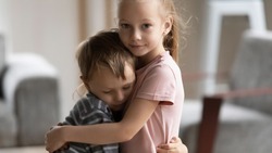 Head shot portrait of little kid girl cuddling smaller brother at home, showing love and care. Compassionate sister comforting soothing upset stressed boy in living room, siblings relations concept.