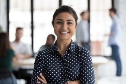Head shot portrait close up smiling confident beautiful Indian businesswoman standing in modern office room with arms crossed, successful executive team leader mentor posing for corporate photo