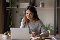 Smiling millennial Caucasian girl in headphones sit at desk look at laptop screen study online, happy smart young woman in earphones take web course or training on computer, distant education concept