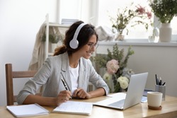 Happy young businesswoman wearing earphones, enjoying studying online, writing notes. Smiling creative designer decorator in eyeglasses looking at computer screen, improving professional skills.