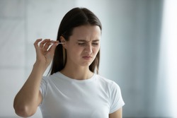 Close up young woman cleaning ears, using cotton bud after shower, feeling pain, beautiful female wearing white t-shirt standing in bathroom, morning routine, personal hygiene concept