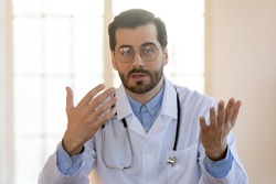 Head shot portrait young doctor wearing glasses and white uniform with stethoscope speaking, consulting patient online, looking at camera, making video call, sitting at table in office