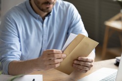 Crop close up of man sit at desk open envelope with paper letter or correspondence at office. Male worker get postal paperwork or notice notification at workplace, receive message or invitation.