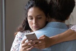 Close up of bored young Caucasian woman hug man texting messaging with lover on smartphone online. Unhappy wife embrace husband cheating on web on cellphone. Family, relationship problem concept.