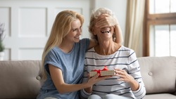 Guess what? Laughing grown up daughter congratulates happy mature mother covering her eyes and giving birthday gift, excited senior grandma receiving surprise on Mothers Day from adult granddaughter