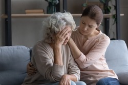Caring grownup daughter comforting frustrated unhappy mature woman, hugging, sitting on couch at home, upset elderly female feeling unwell, touching forehead, suffering from migraine or head ache