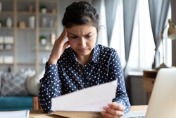 Indian woman sit at desk hold document read paper letter feels disappointed shocked by bad news. Financial trouble bank debt notice, medical test results health problems, huge domestic bills concept