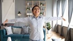 Lively Indian girl having wide smile standing with arms stretched eyes closed enjoy weekend at new house feels free from life troubles, sunlight illuminates cozy room. Welcoming new happy day concept
