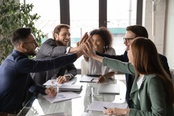 Overjoyed young mixed race business people sitting at table, joining hands in air, showing group unity, celebrating successful teamwork or professional achievement, raising team spirit at meeting.