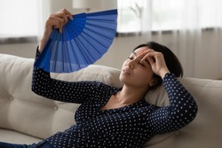 Unwell young Asian woman sit rest on sofa at home wave with hand fan suffer from hot weather heatstroke. Unhealthy Vietnamese female use waver struggle with hormonal imbalance or AC lack indoors.