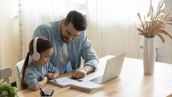 Caring father helping little daughter with school homework, sitting at table at home, child schoolgirl wearing headphones studying online, using laptop, dad checking tasks, homeschooling concept