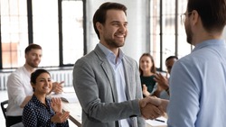 Happy motivated millennial man intern is being hired on regular job employed to international company, loyal young male manager handshaking with leader employer on formal meeting with corporate team