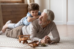 Overjoyed mature grandfather playing with adorable grandson at home, older man lying on warm floor with cute little boy grandchild on back, family spending leisure time together, having fun
