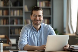 Good-looking millennial office employee in glasses sitting at desk in front of laptop smiling looking at camera. Successful worker, career advance and opportunity, owner of prosperous business concept