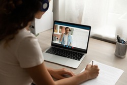 Back view focused young woman in headphones looking at computer screen, holding video call online private lesson with male teacher native speaker, learning foreign language distantly at home.