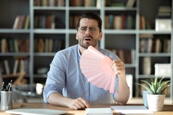 Man office manager sit at workplace desk hold pink handheld fan waving it to reduce unbearable hot weather suffers from high temperature indoor at summer workday in modern room without air conditioner