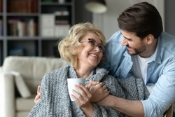 Smiling mature woman holding cup, hugging with adult son close up, enjoying leisure time, happy elderly mother wearing warm scarf drinking hot tea or coffee, family spending weekend together at home