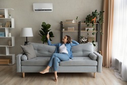 Happy young Caucasian woman relax on couch in living room turn on air conditioner with remote controller. Smiling female rest on sofa at home breathe fresh air from ac electronic condition device.