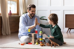 Loving young Caucasian dad sit on floor play with small son give high five celebrate success. Happy caring father engaged in activity game build with blocks bricks with little boy child together.