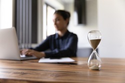 Busy young mixed race woman working studying by laptop on blurred background doing urgent job taking exam. Focus on close up sand glass posed on desk at home office. Measuring time. Deadline is coming