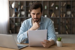 Pensive young Caucasian man sit at desk at home work on laptop read paper document. Thoughtful millennial male distracted from computer job consider post paperwork or letter correspondence news.