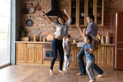 Barefoot dances. Overjoyed millennial family with little daughter son having fun listening to music dancing. Active mom dad and two junior school age kids jump at kitchen celebrate moving to new home