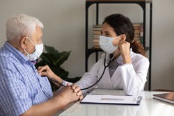 Caring female nurse in medical facemask listen to elderly patient heart with stethoscope in hospital. Woman GP or doctor in facial mask do checkup examine mature man client on consultation in clinic.
