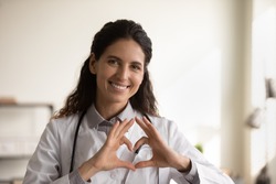 Close up portrait of smiling young Caucasian female nurse or GP in white medical uniform show heart love hand gesture. Happy woman doctor show support and care to patients or client in hospital.