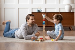 Caring young Caucasian father and small son sit on warm floor at home engaged in funny game together. Loving dad and little boy child have fun play build construct with wooden blocks bricks.