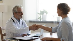 Side view smiling middle aged professional physician in white coat and eyeglasses shaking hands with happy young woman, welcoming at checkup meeting or getting acquainted with new patient indoors.
