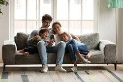 Happy bonding family with two adorable small kids looking at cellphone screen, laughing at funny video or photos in social networks, holding distant video call with grandparents, having fun at home.
